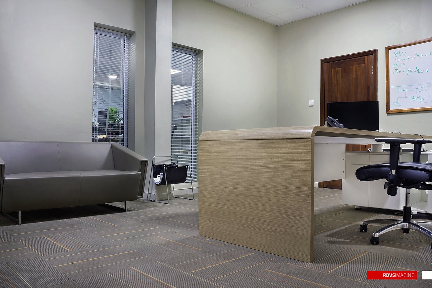 Photograph of a small office: pale wood desk with a computer, black task chair, grey sofa against a white wall, grey tiled floor, timber door, framed board and a window with blinds looking onto another building.