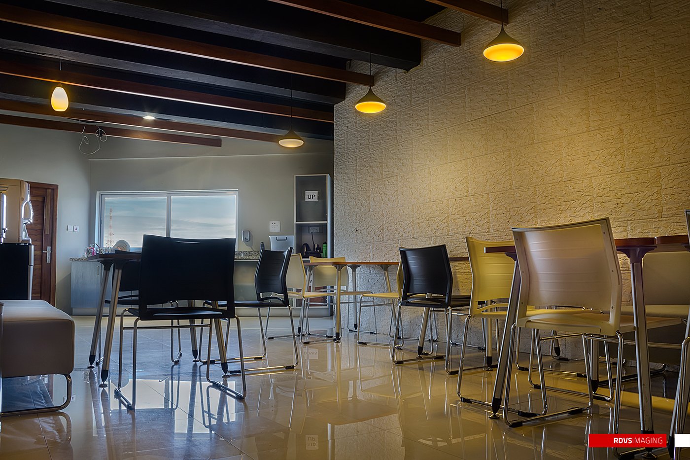 Photograph of the break area: dark ceiling beams with yellow pendant lamps, high tables with black, yellow and tan stools, white painted brick, a sofa at left and a glossy floor underfoot.