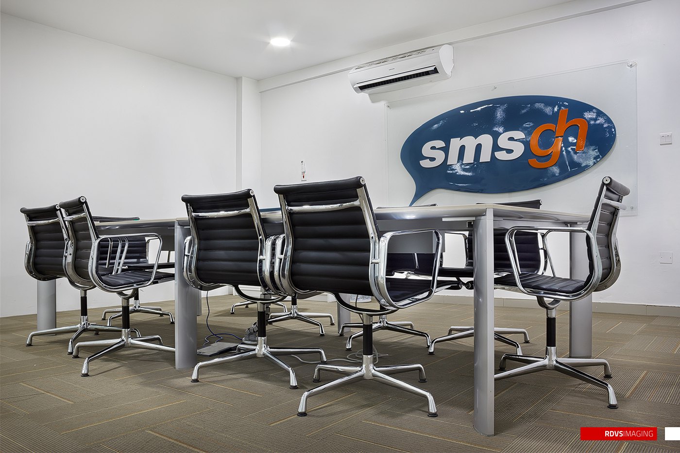 Visualization of a meeting room: the sms gh speech-bubble logo mounted on a white wall, grey tiled floor, a pale table ringed by black and chrome chairs, air-conditioning unit above and a downlit white ceiling.
