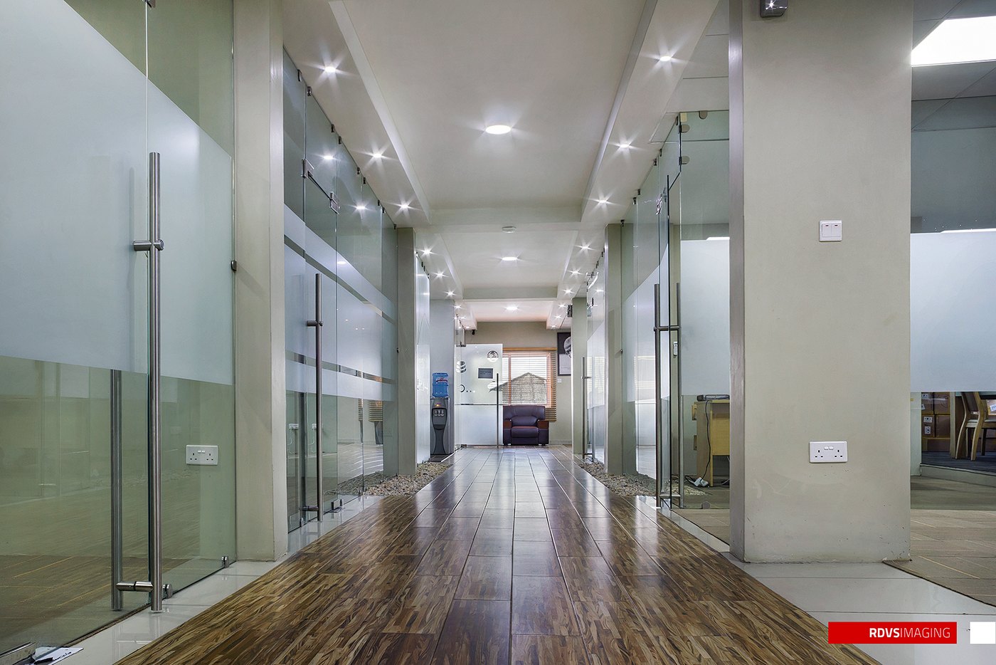 Photograph of a completed corridor: glass-walled offices with frosted horizontal strips, polished dark timber floor reflecting the downlights, an exposed concrete column, white ceilings and a glimpse of the workspace at the far end.