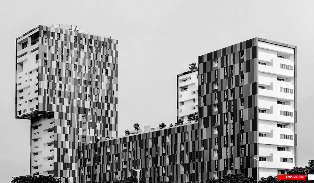 A black and white view across the tops of three residential volumes whose facades are clad in an irregular pixel mosaic of pale, mid and dark grey panels. The tallest block stands at the left with a stack of white recessed balconies down its outer flank and an open, unglazed storey at its crown; a second tower rises at the right with the same checkered cladding and white balcony flank; a low block joining them carries small trees along its roofline. The sky is flat featureless white, a thin treeline closes the bottom of the frame, and the only colour left in the picture is the red rectangular RDVSIMAGING badge in the bottom-right corner.