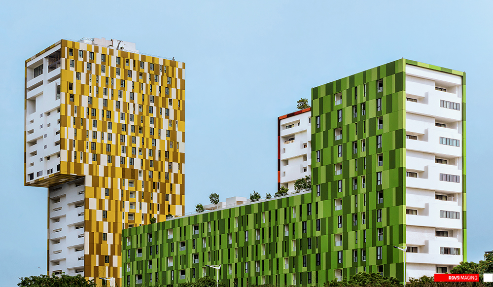 The same daylight view printed in full colour: the tall tower at the left is clad in an irregular mosaic of yellow, gold and cream rectangular panels with a flank of plain white recessed balconies, and the tower at the right and the low block joining them are clad in the same mosaic in greens. The left tower has an open, unglazed storey and a small white rooftop structure at its crown; young trees and a white box sit along the podium roof between the towers. The sky is a flat pale blue-grey, a treeline closes the bottom of the frame, and a red RDVSIMAGING badge with a small white square tag sits in the bottom-right corner.