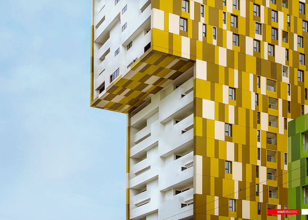 A close daylight crop of the yellow tower, taken at the level where an upper volume oversails the floors below it. The checkered yellow, gold and cream cladding wraps over the underside of the overhang, so the pattern runs diagonally across the soffit, while beneath it the white balconies step back in a stack of deep horizontal openings with narrow windows set into the recesses. Windows pierce the tiled face irregularly down the right side of the frame, the pale green edge of the neighbouring block just enters at the bottom right, and the sky at the left is clear pale blue. Red RDVSIMAGING badge and white tag, bottom right.