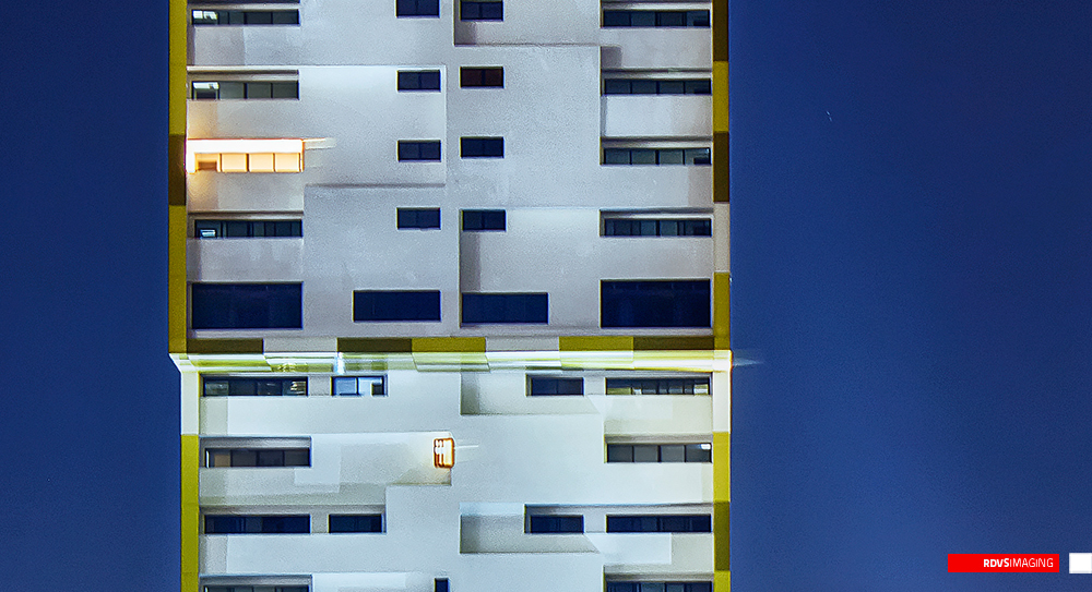 A night crop across the waist of the tall tower, landscape rather than the verticals around it. The white facade with its staggered recessed balconies and dark windows fills most of the frame, one window lit warm orange at the upper left and a smaller one lower down, and the yellow cladding reads as a thin lit band at the left edge. A horizontal streak of white light smears right across the middle of the image at the joint between the two volumes, the trail of a moving light source during a thirty-second exposure, and the sky behind the building is a deep even blue. Red RDVSIMAGING badge and white tag, bottom right.