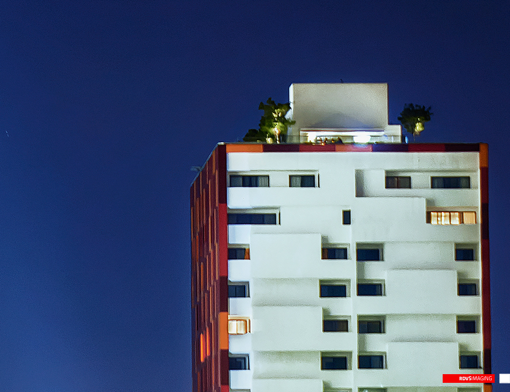 The crown of the red tower at night: a white penthouse box and lift overrun sit on the roofline with planted trees lit from below, and the top storeys beneath are white with staggered balconies, one window glowing warm orange at the right and another at the lower left. The tower&rsquo;s return face along the left is clad in the same pixel mosaic, reading here as deep red and orange in the low light, and the sky is a solid dark blue with a faint streak near the left edge. Red RDVSIMAGING badge and white tag, bottom right.