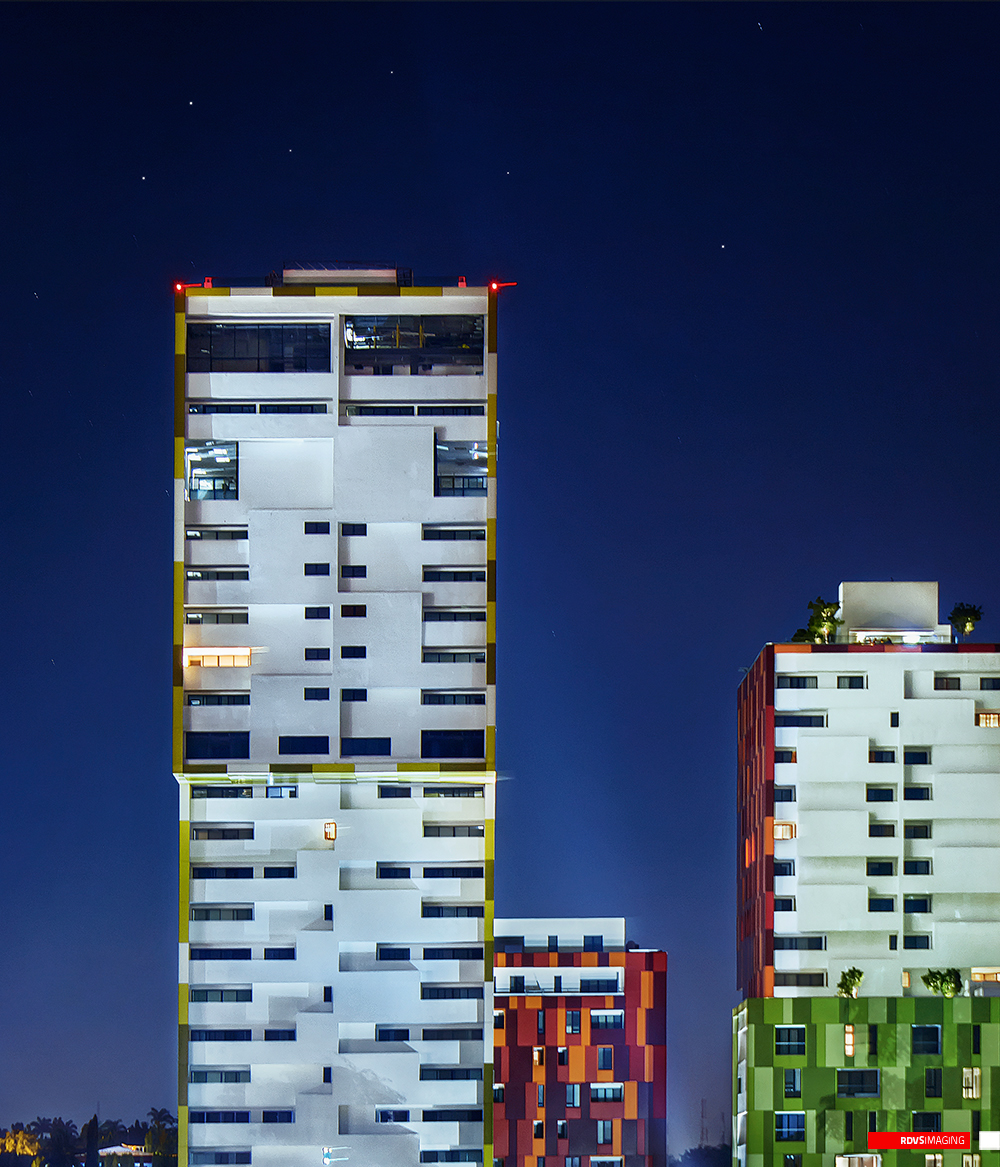 A night crop of two towers against a dark blue sky pricked with star points. The left tower, edged in yellow cladding with white staggered balconies, is topped by two red obstruction lights and an open unglazed storey, with a few lit windows and a warm orange one lower down; the right tower is clad in red and orange panels with a white crown and a planted, floodlit roof. Between and below them the tops of two more pixel-clad blocks appear, one red and one green, and palms and low lit buildings run along the horizon at the bottom left. Red RDVSIMAGING badge and white tag, bottom right.