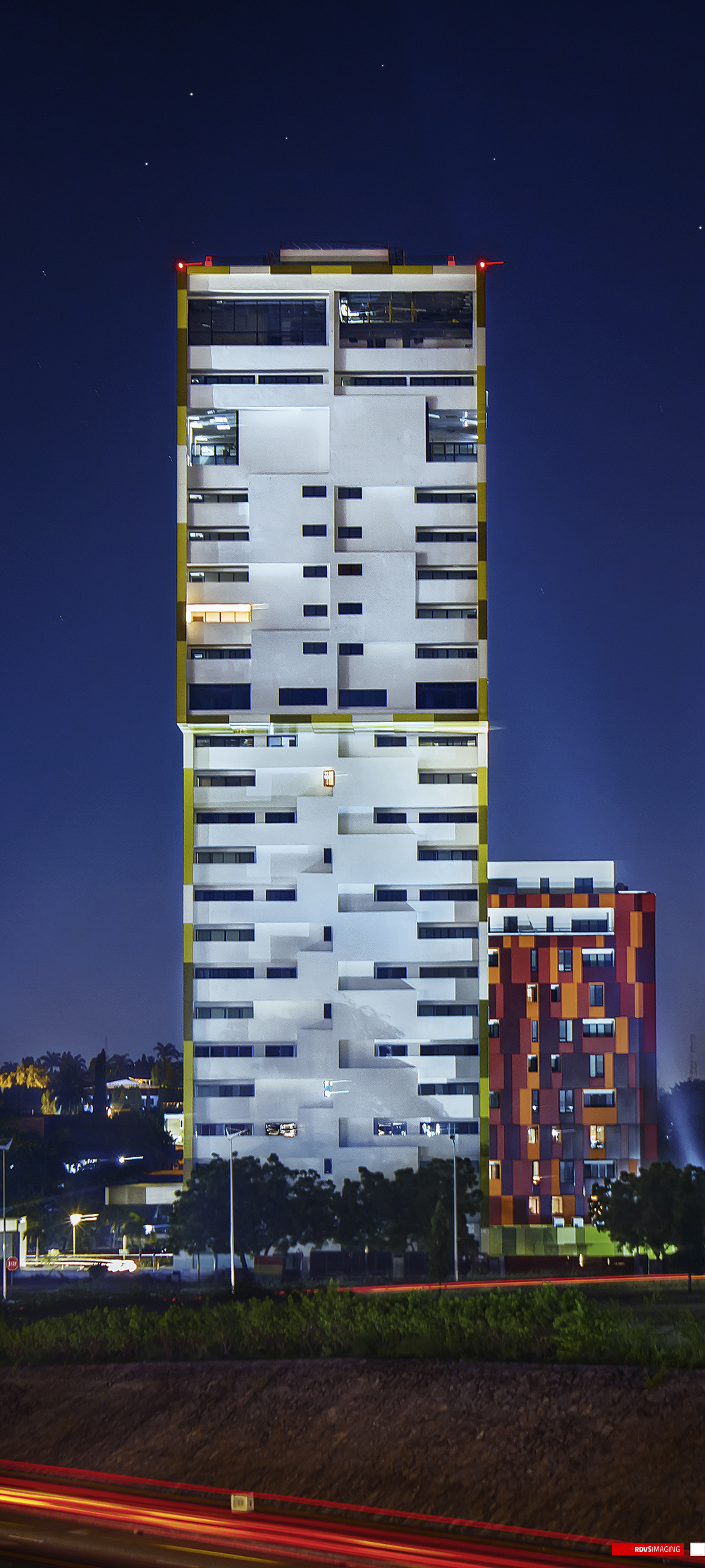 The tallest plate, the whole night scene cropped narrow and vertical. The yellow-edged tower stands full height through the centre, its crown lit by two red obstruction lights above an open unglazed storey, its white balconies broken by a few lit windows and by a horizontal light trail at the joint between its two volumes; the red and orange pixel-clad tower stands shorter at the right with scattered lit windows. Below them a road runs across the frame streaked red and white by traffic, with street lamps and trees at the base of the towers, palms and low lit buildings at the left, and a dark sky holding still stars above. Red RDVSIMAGING badge and white tag, bottom right.