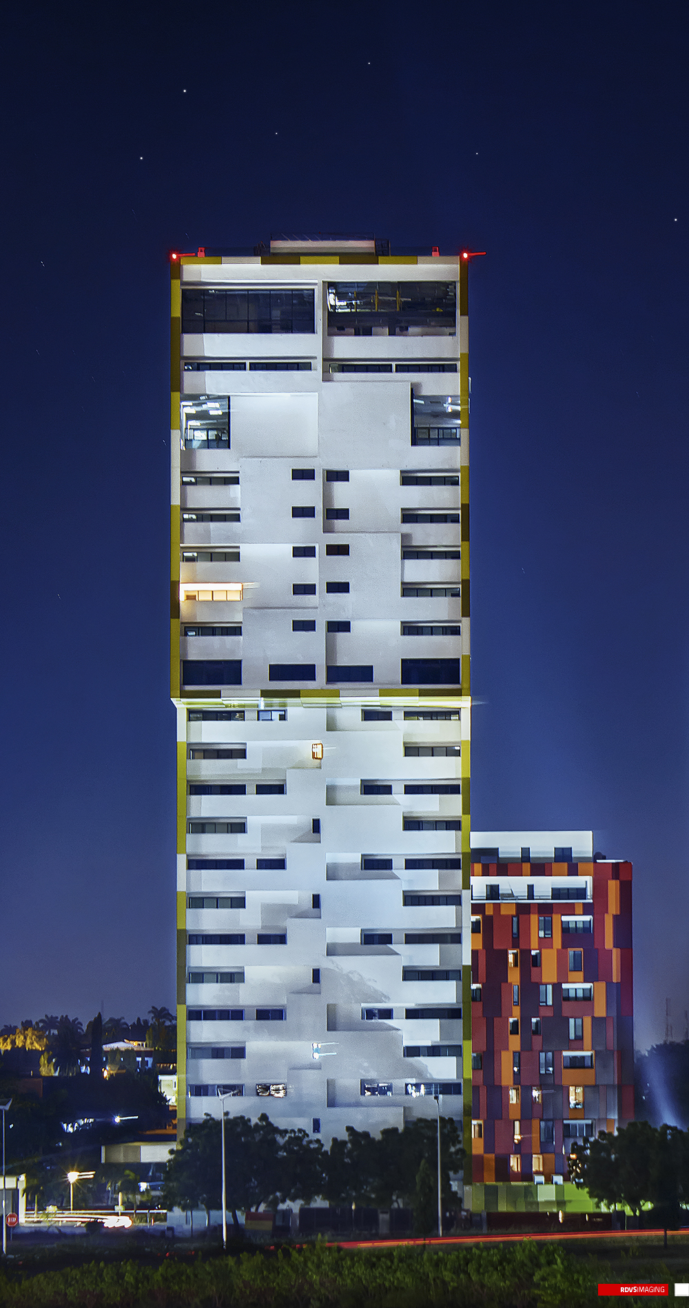The same night view a little tighter: the yellow tower fills the left two-thirds from its red obstruction lights down through its lower floors, the red and orange pixel-clad tower sits at the right with lit windows scattered across its facade, and only the treetops and a thin band of lit road survive of the foreground. Star points are visible in the deep blue sky, a light trail smears across the waist of the taller tower and another streaks along the road at the bottom. Red RDVSIMAGING badge and white tag, bottom right.