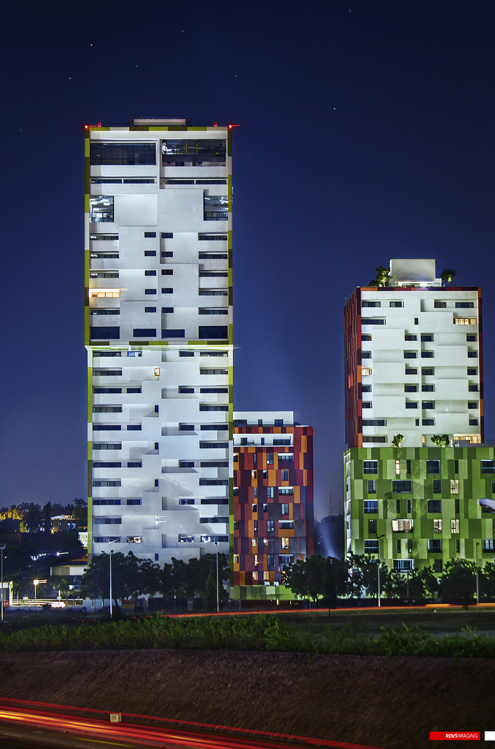 The broadest night crop, taking in three towers at once: the tall yellow-edged block at the left with its open crown and red obstruction lights, a shorter red and orange pixel-clad block in the middle distance, and the red tower at the right rising above a green pixel-clad base with its planted roof floodlit from below. Street lamps, trees and a low lit wall run along the middle ground, red and white light trails streak the road across the bottom of the frame, and stars are scattered through the dark blue sky. Red RDVSIMAGING badge and white tag, bottom right.