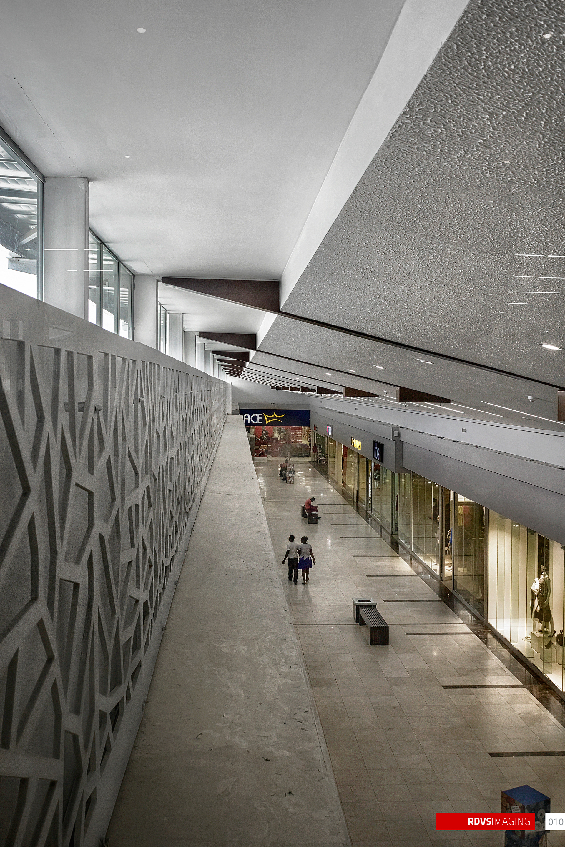 A mall interior looking down an upper-level walkway, shot portrait so the corridor runs away from the camera and the ceiling fills the top third. Along the left runs a tall solid parapet whose face is a decorative screen cut with a repeating lattice of elongated hexagons and star shapes, and above it a band of clerestory glazing with visible steel framing lets in flat daylight. On the right is a two-storey retail frontage: a blue fascia sign ending in the letters ACE with a crown device above it, a small black square sign beside it, and lit shopfronts at ground level with a mannequin in the nearest window. The ceiling is a broad angled band of sprayed textured soffit studded with recessed downlights, stepping down toward the far end. The floor is large pale tiles with darker inset strips, a slatted bench sits at the right, two people walk away from the camera in the middle distance and one more is seated further along. A red RDVSIMAGING badge and a small white tag numbered 010 sit in the bottom-right corner.