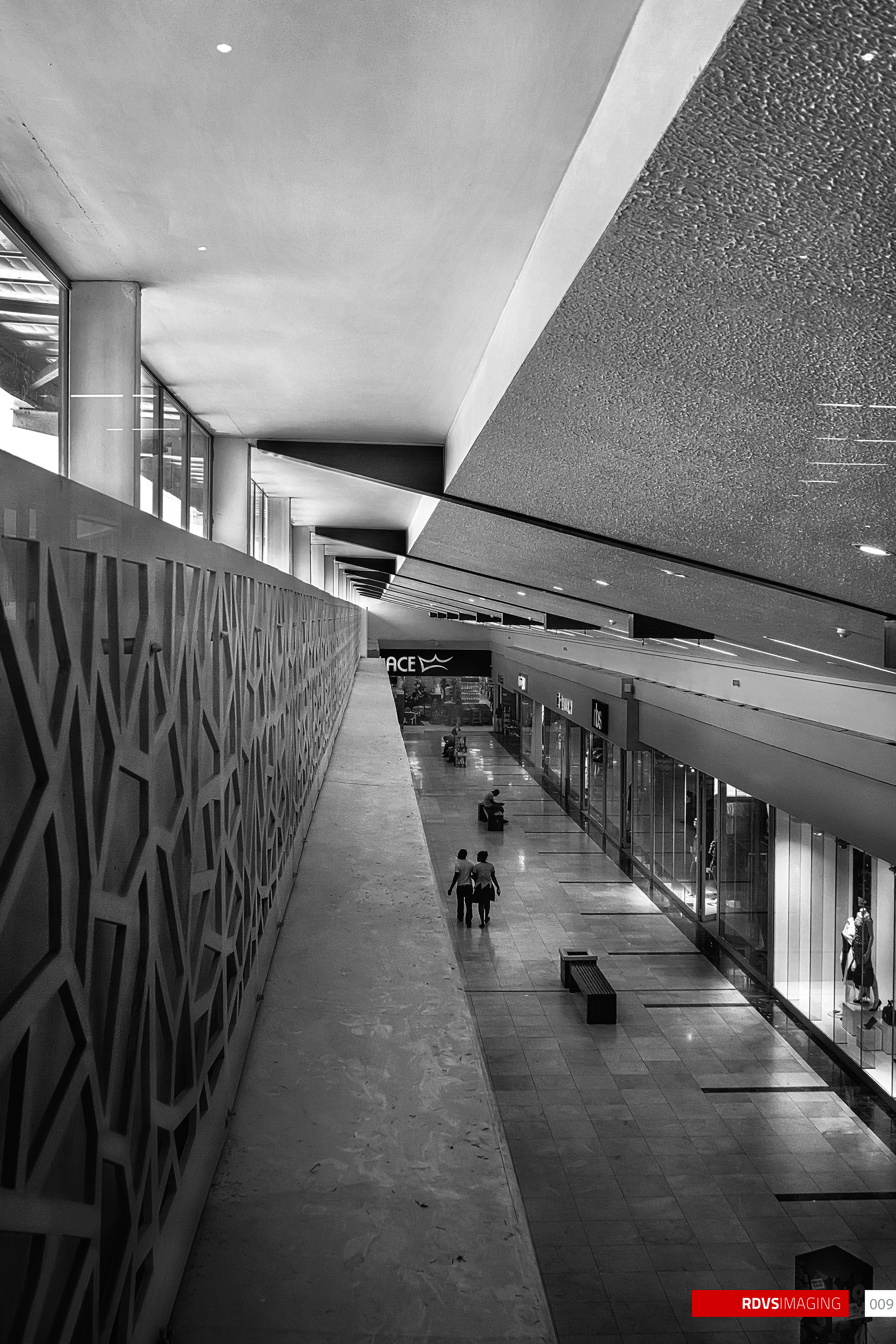 The same view, same framing and the same people in the same positions, printed in black and white: the lattice screen at the left goes from pale grey to near-black in its cut openings, the textured soffit becomes a field of fine grain, the blue fascia sign reads as dark with light lettering, and the daylight through the clerestory blows out to pure white. A red RDVSIMAGING badge and a small white tag numbered 009 sit in the bottom-right corner.
